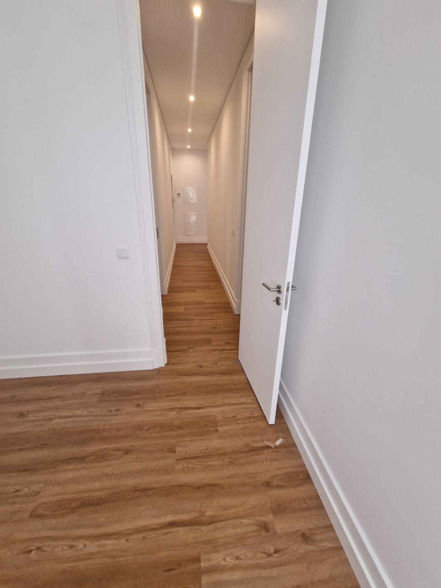 Corredor estreito com piso de madeira clara, paredes brancas e uma porta branca parcialmente aberta.