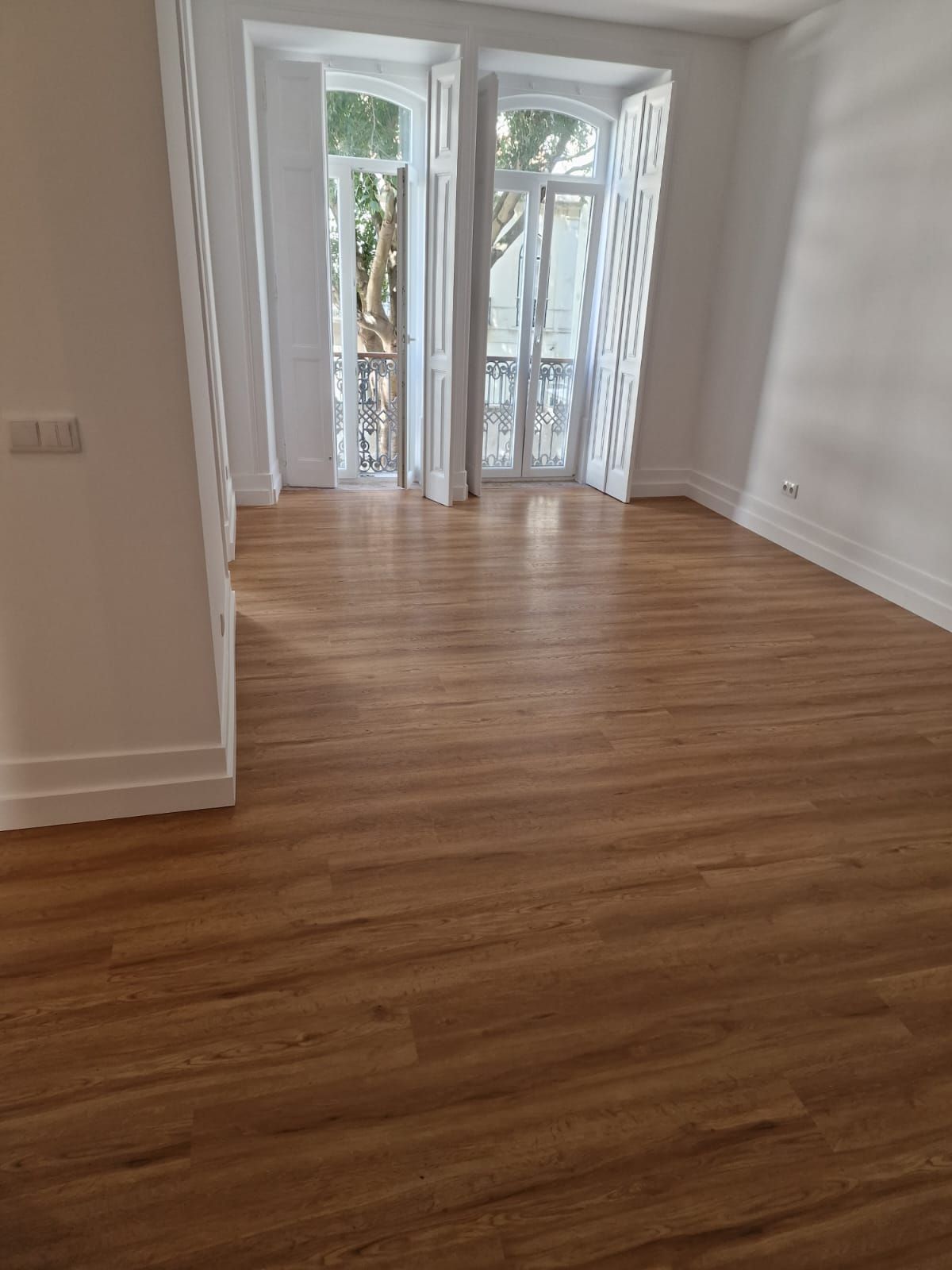 Quarto vazio com piso de madeira e uma grande janela com cortinas brancas.