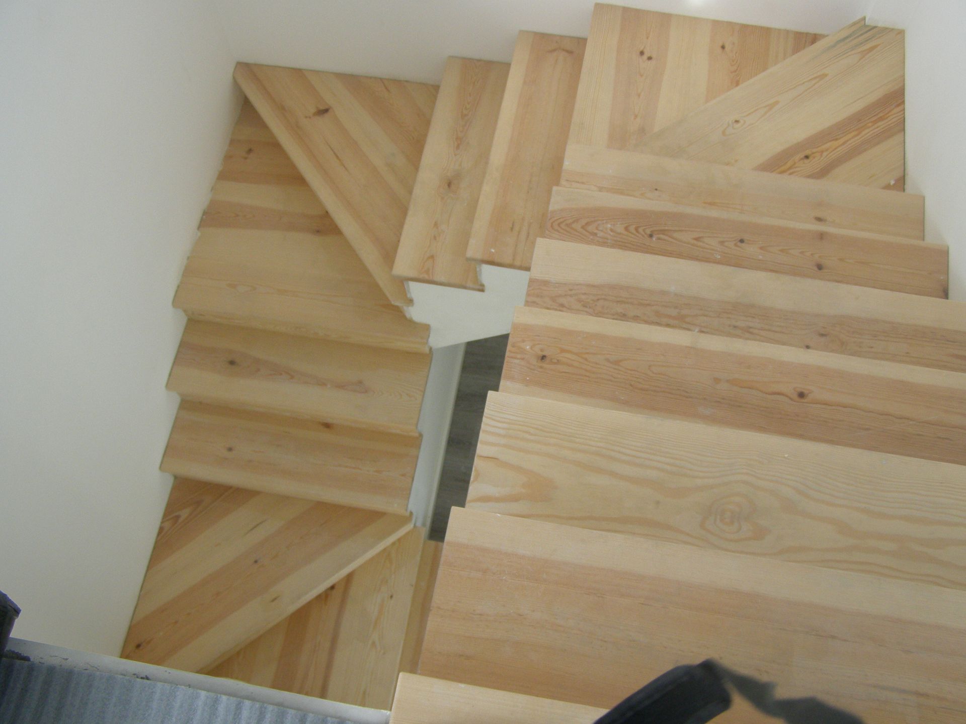 Escadas de madeira, de cor clara com padrão chevron, descem de uma parede branca.