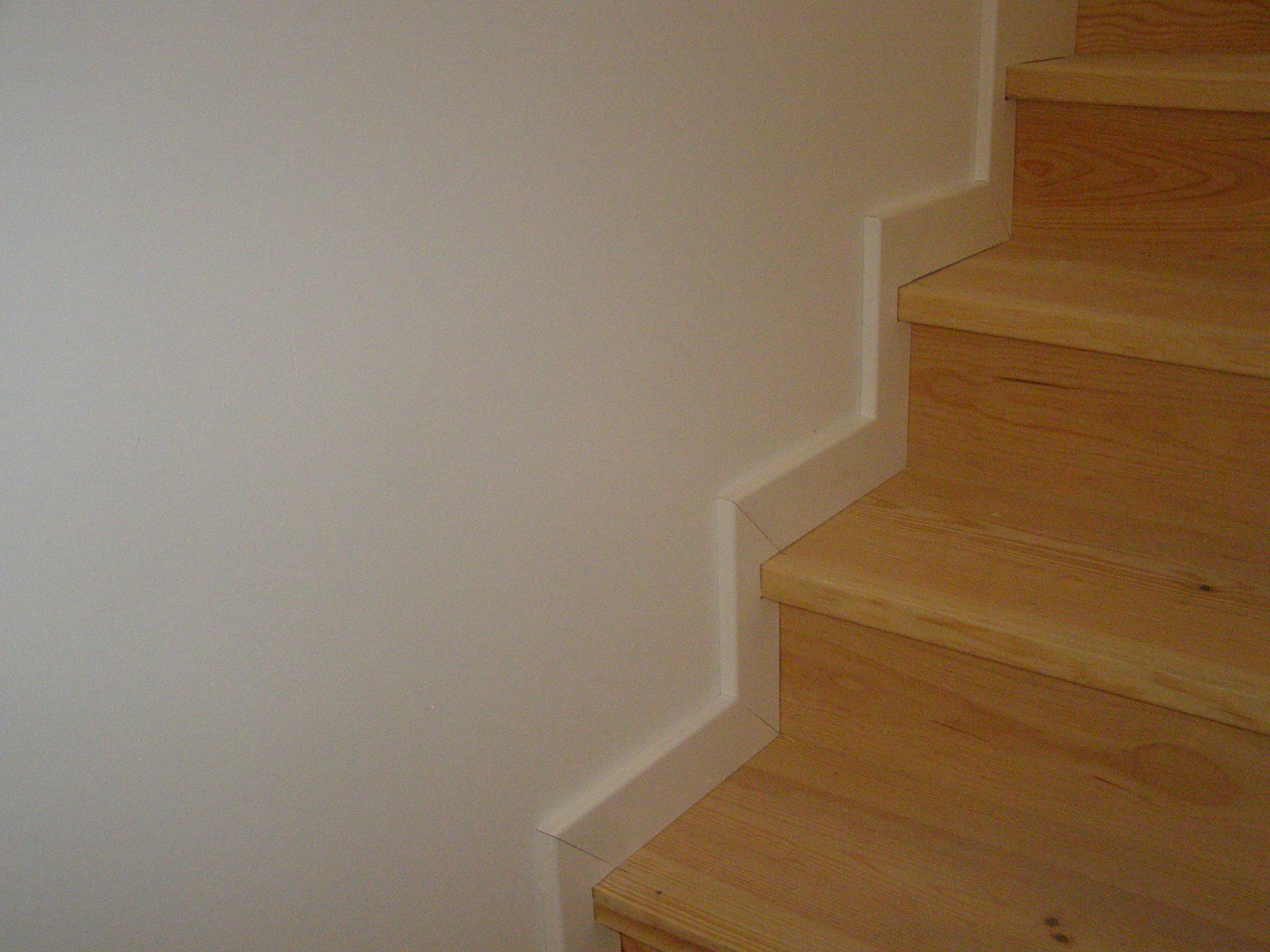 Escada de madeira com parede branca; uma moldura em forma de degrau percorre a parede e a escada.