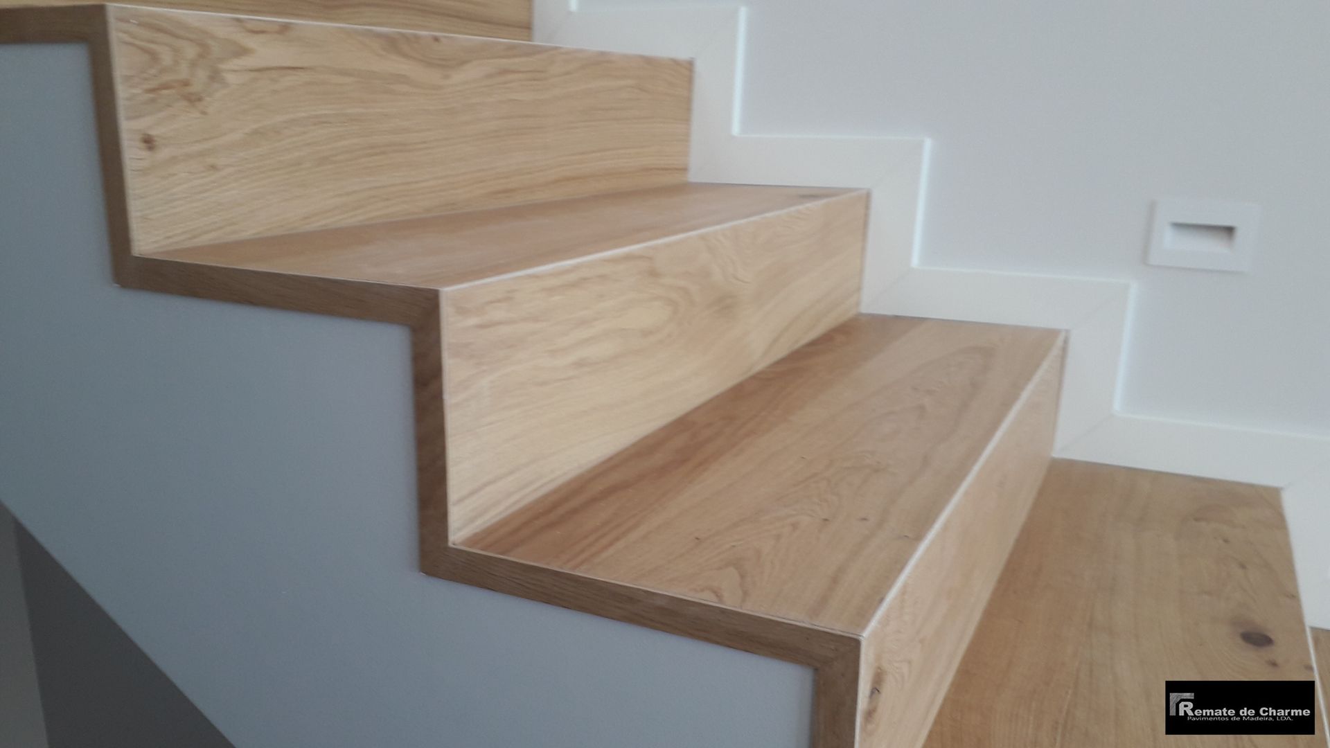 Escada de madeira com acabamento em carvalho claro, descendo em direção a uma parede branca e cinza.