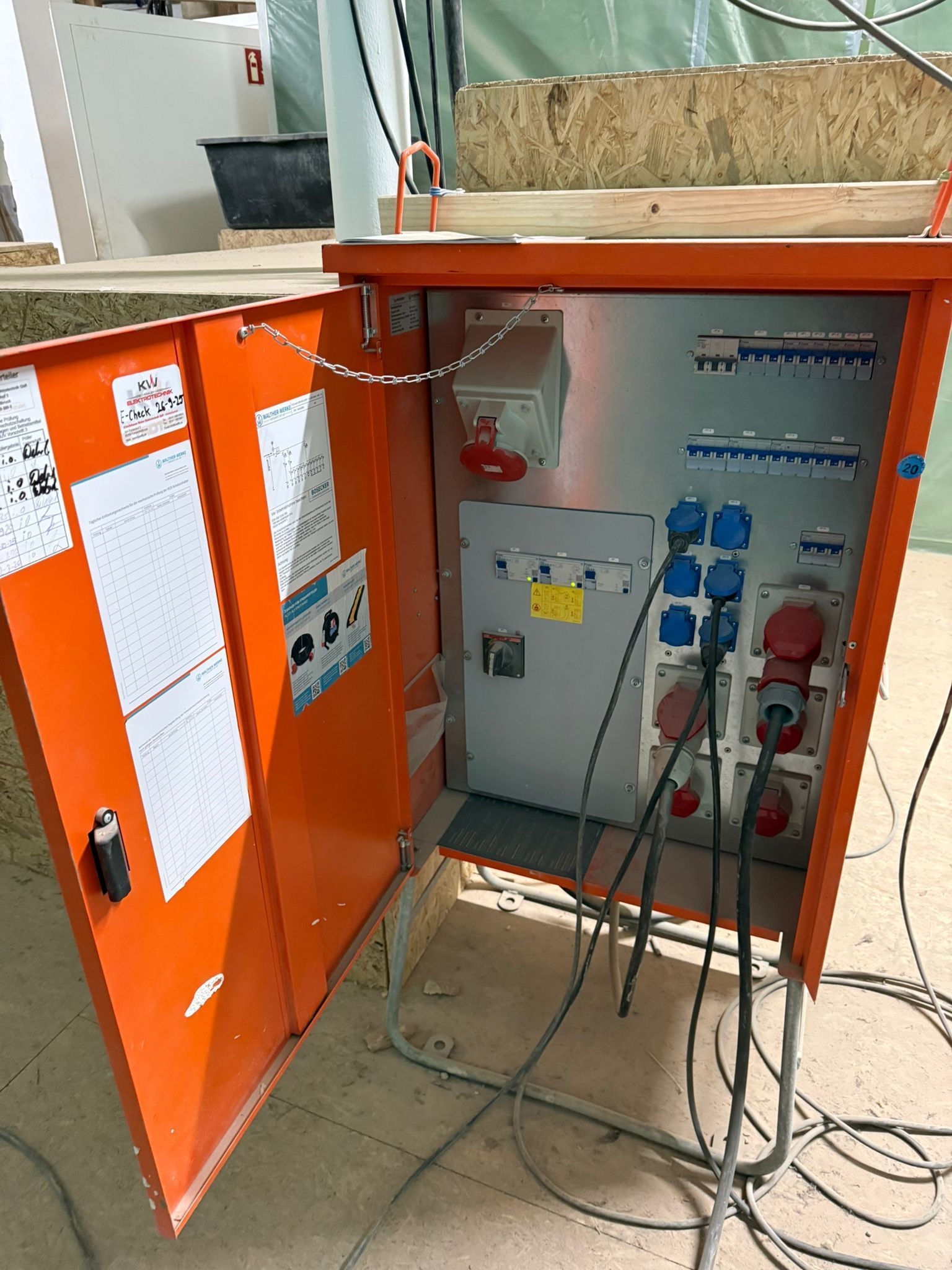 Ein orangefarbener, tragbarer Stromverteilerkasten mit verschiedenen Steckdosen und Schaltern steht auf einer Baustelle.