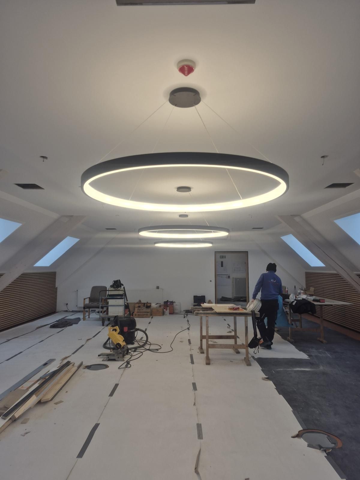 Innenaufnahme eines im Bau befindlichen Dachbodens, von dessen Decke drei große runde Deckenleuchten hängen.