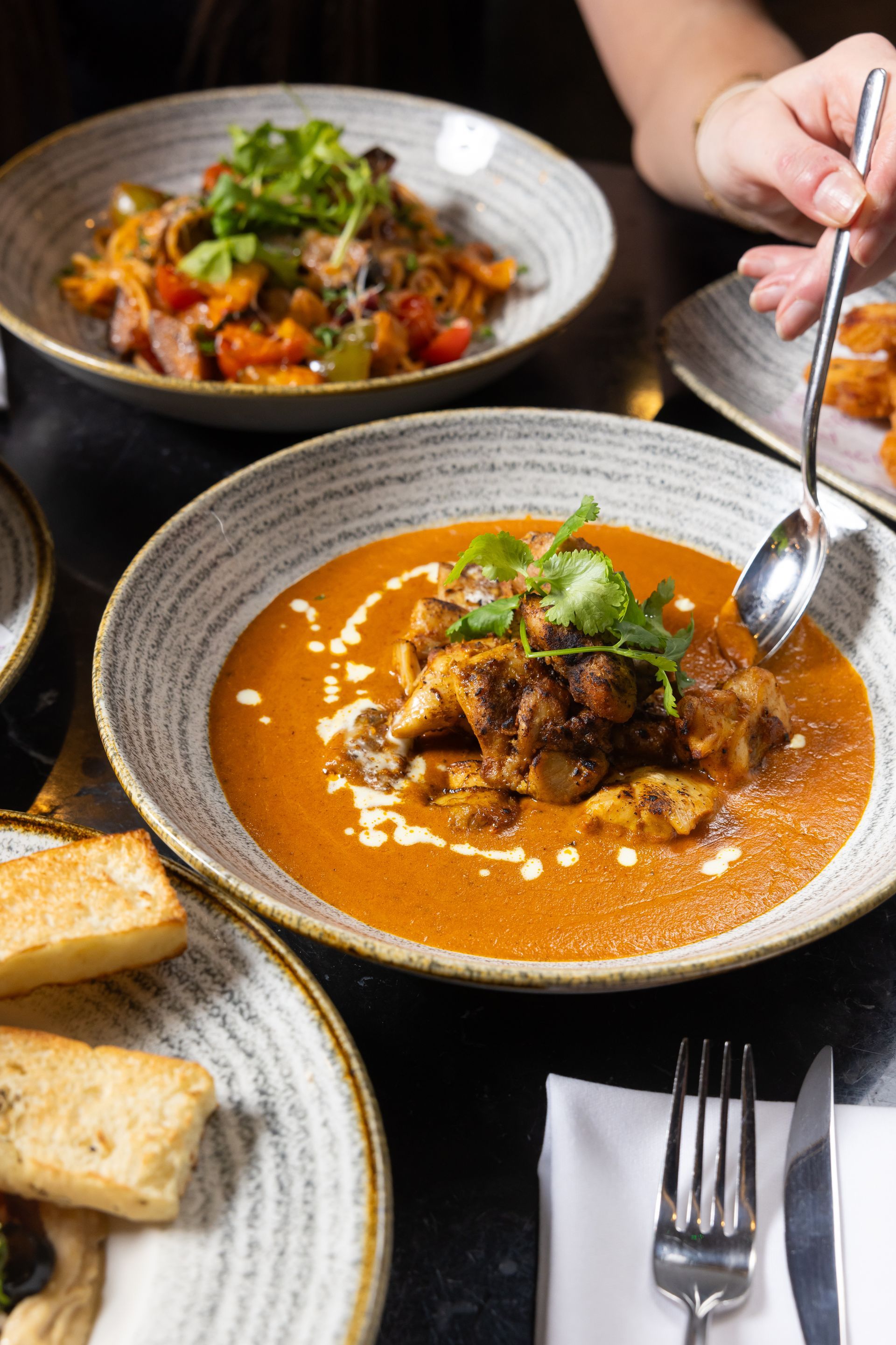 A bowl of Indian butter chicken with garnishes; someone uses a spoon.