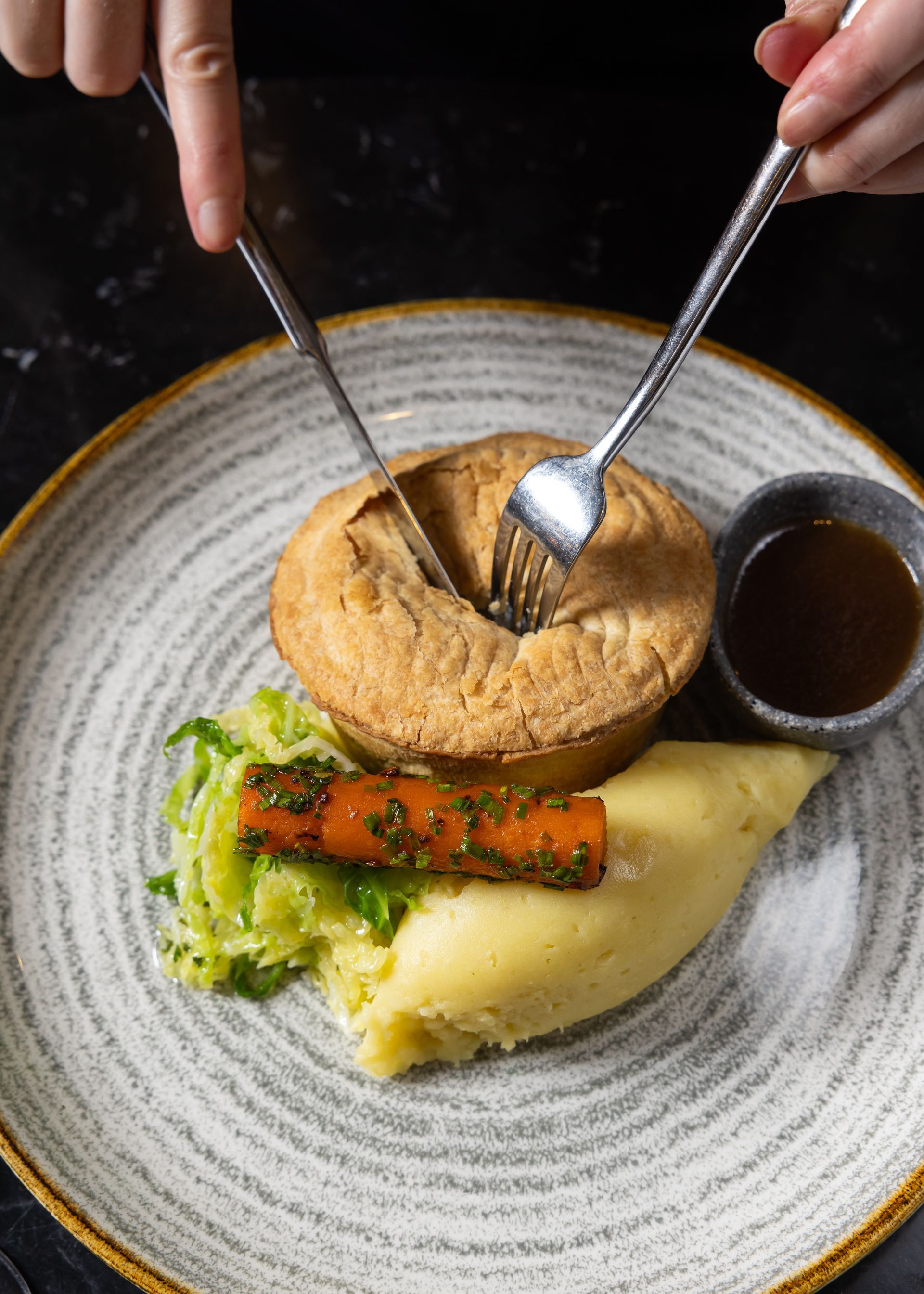 Hands cutting open a meat pie on a plate with mashed potatoes, a carrot, gravy, and greens.