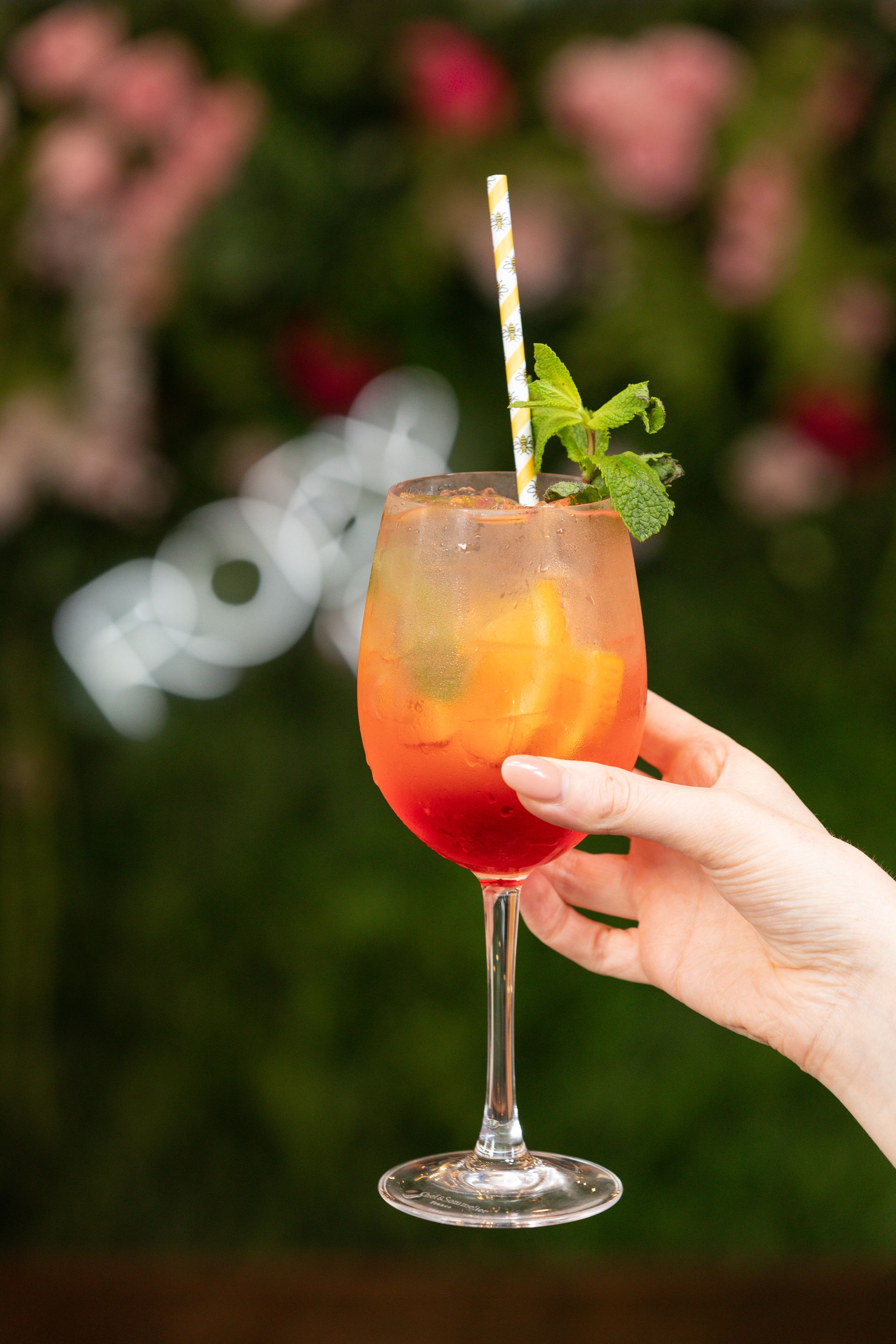 Hand holding a tall cocktail in a stemmed glass. Orange and red gradient drink with mint and straw, against a floral background.