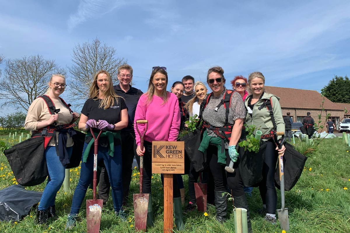 Group of people smiling, planting trees in a sunny field. A sign reads 