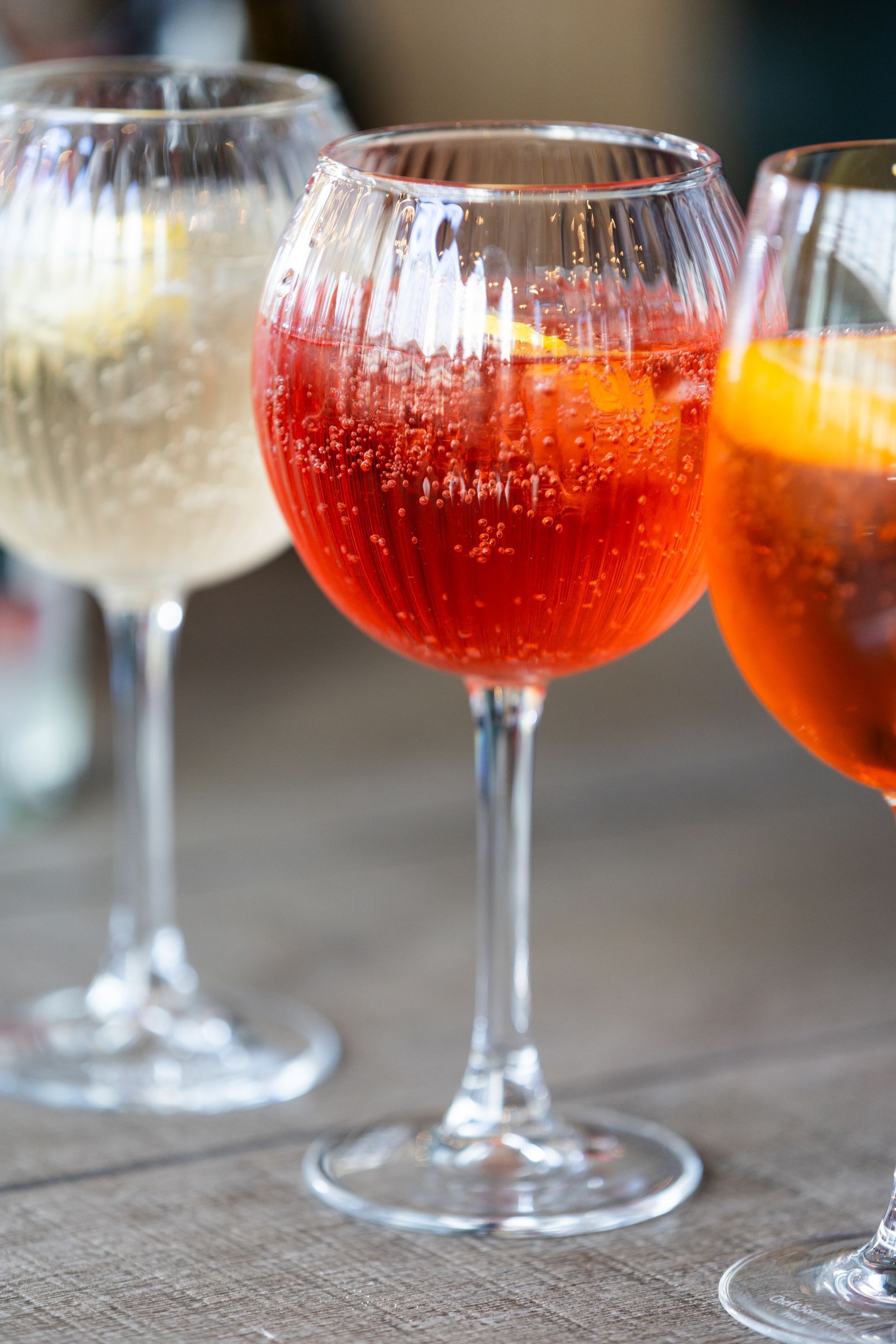 Three stemmed glasses with colorful cocktails: red, orange, and white, bubbles visible, on a table.