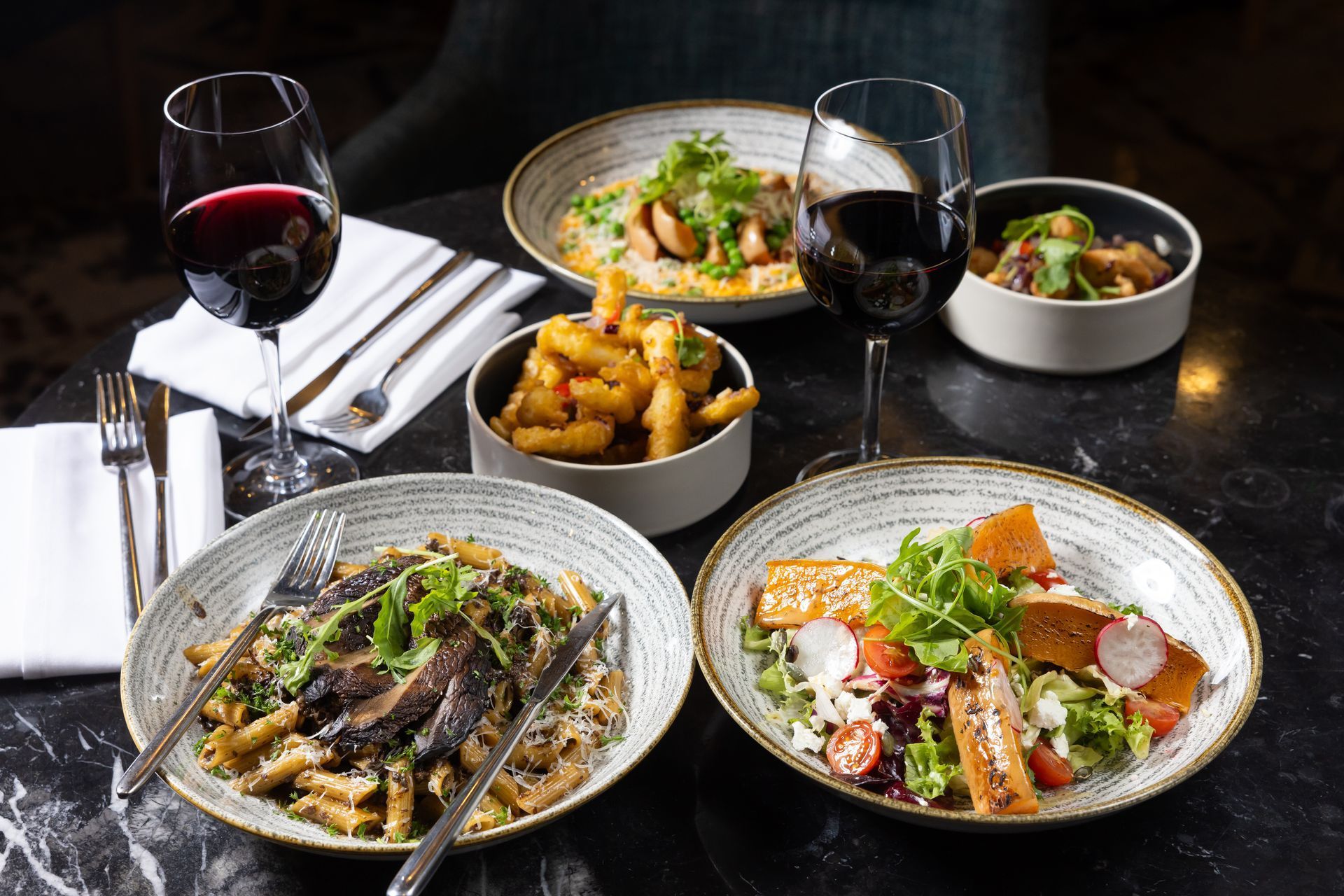 Table set with pasta, salad, fries, and wine glasses, with red wine, in a restaurant.