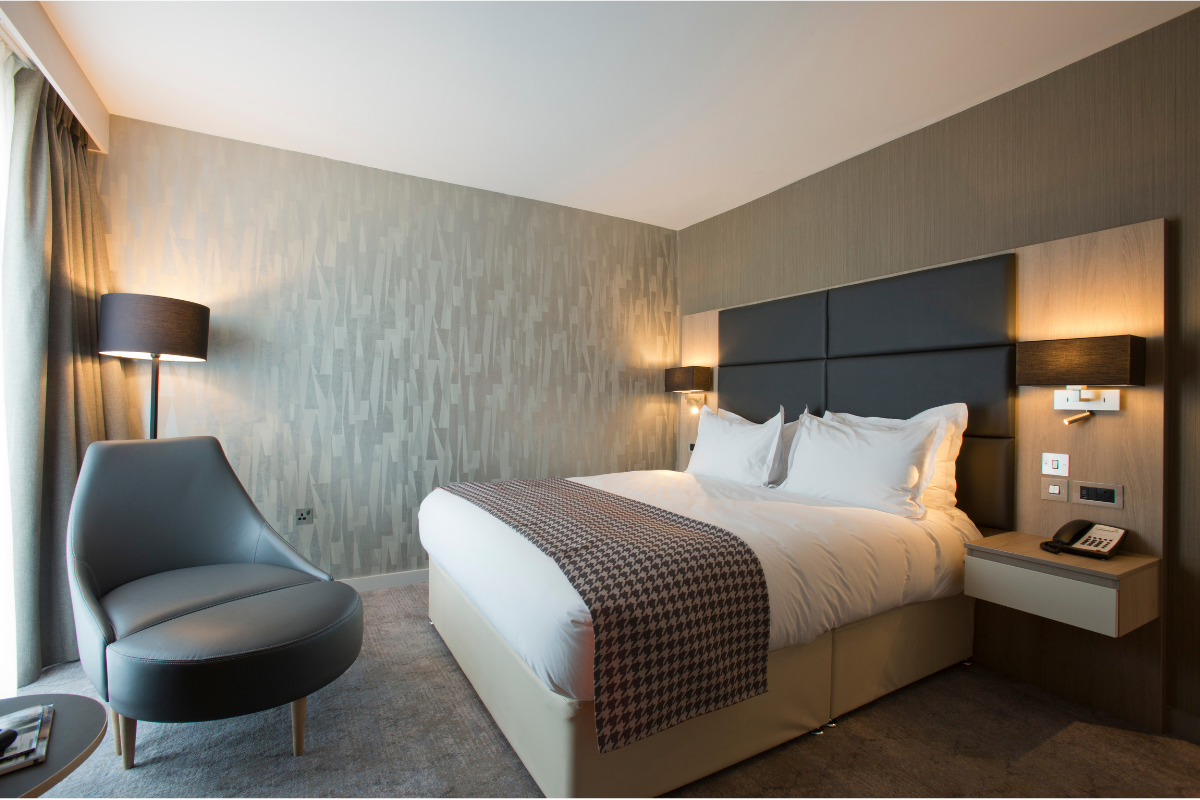 Hotel bedroom with a bed, armchair, and lamp. Neutral tones dominate the space.