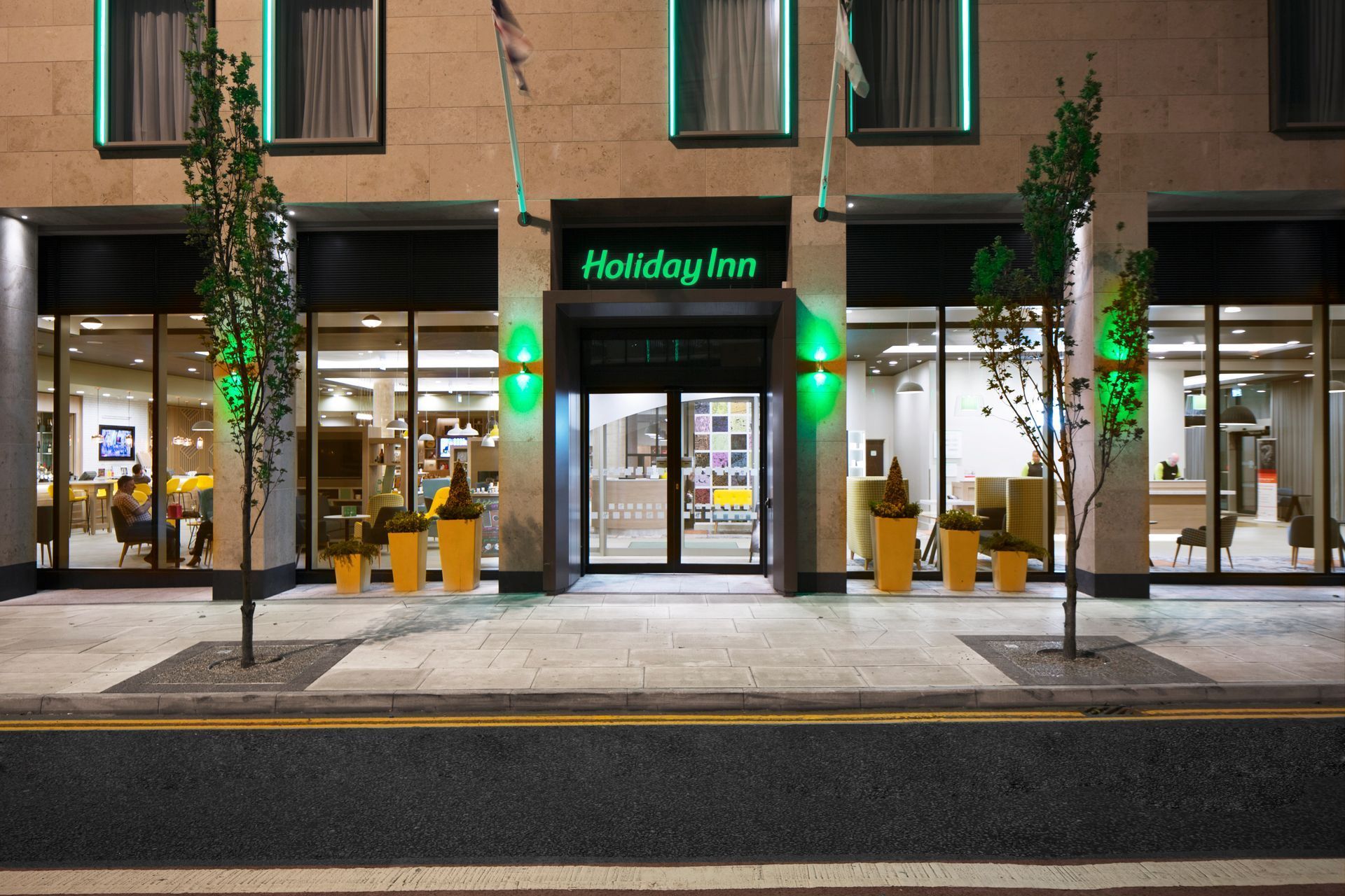 Holiday Inn hotel entrance at night with green illuminated sign and potted plants.