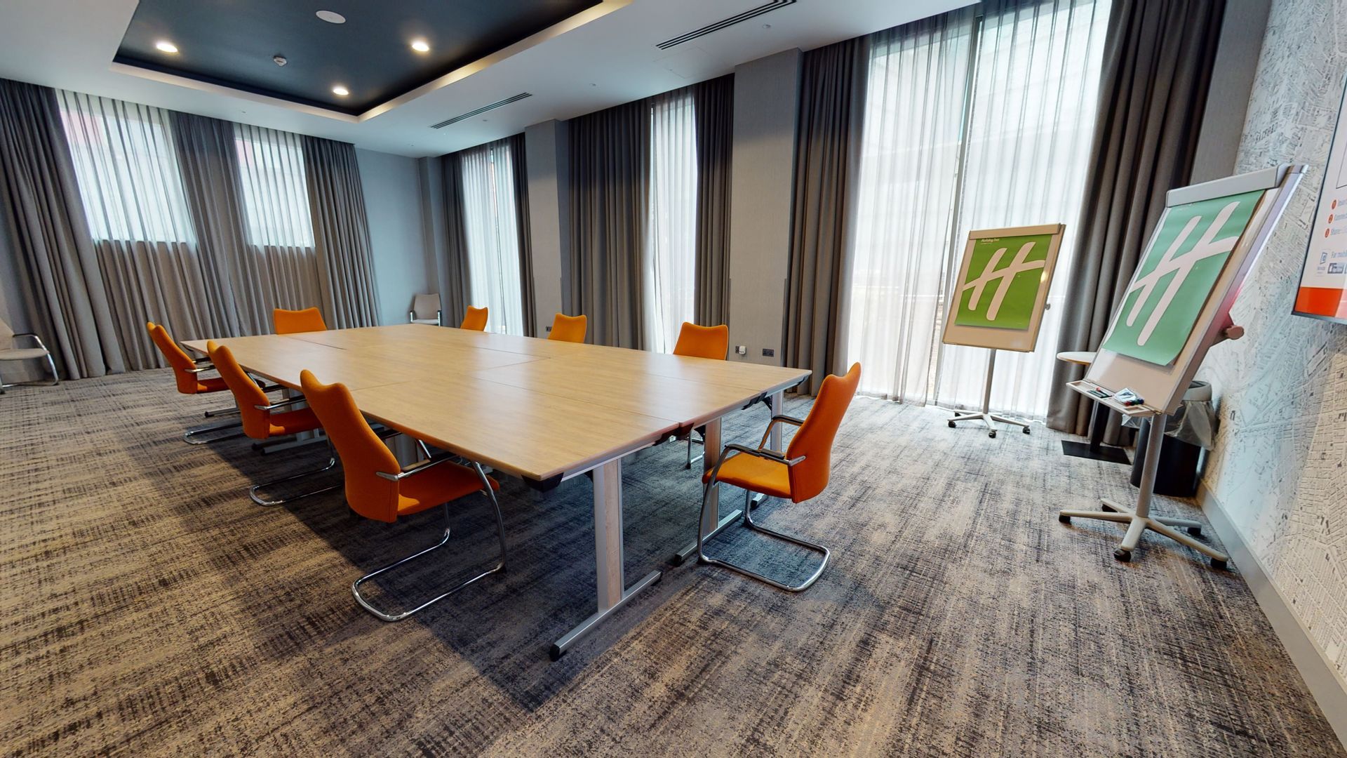 Meeting room with a long table and orange chairs. Holiday Inn sign. Grey carpet and curtains.