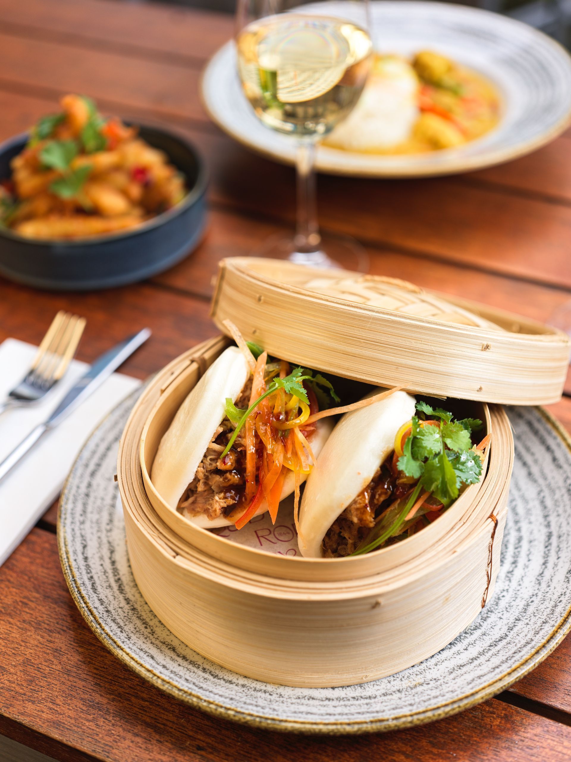 Bao buns in bamboo steamer, other dishes, wine glass, and silverware on wood table.