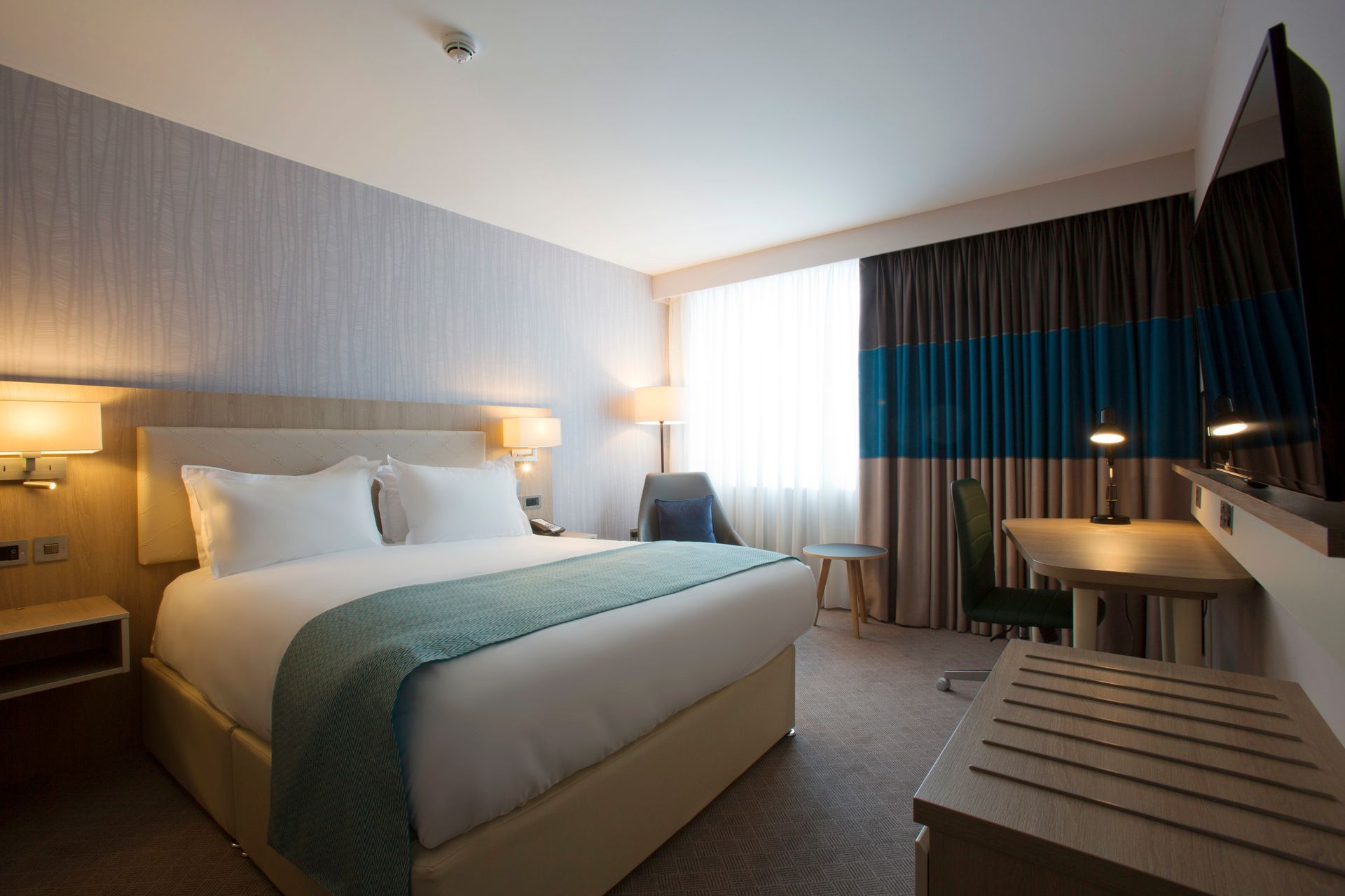 Hotel room: bed with white linens, desk, and large window with blue and gray curtains.