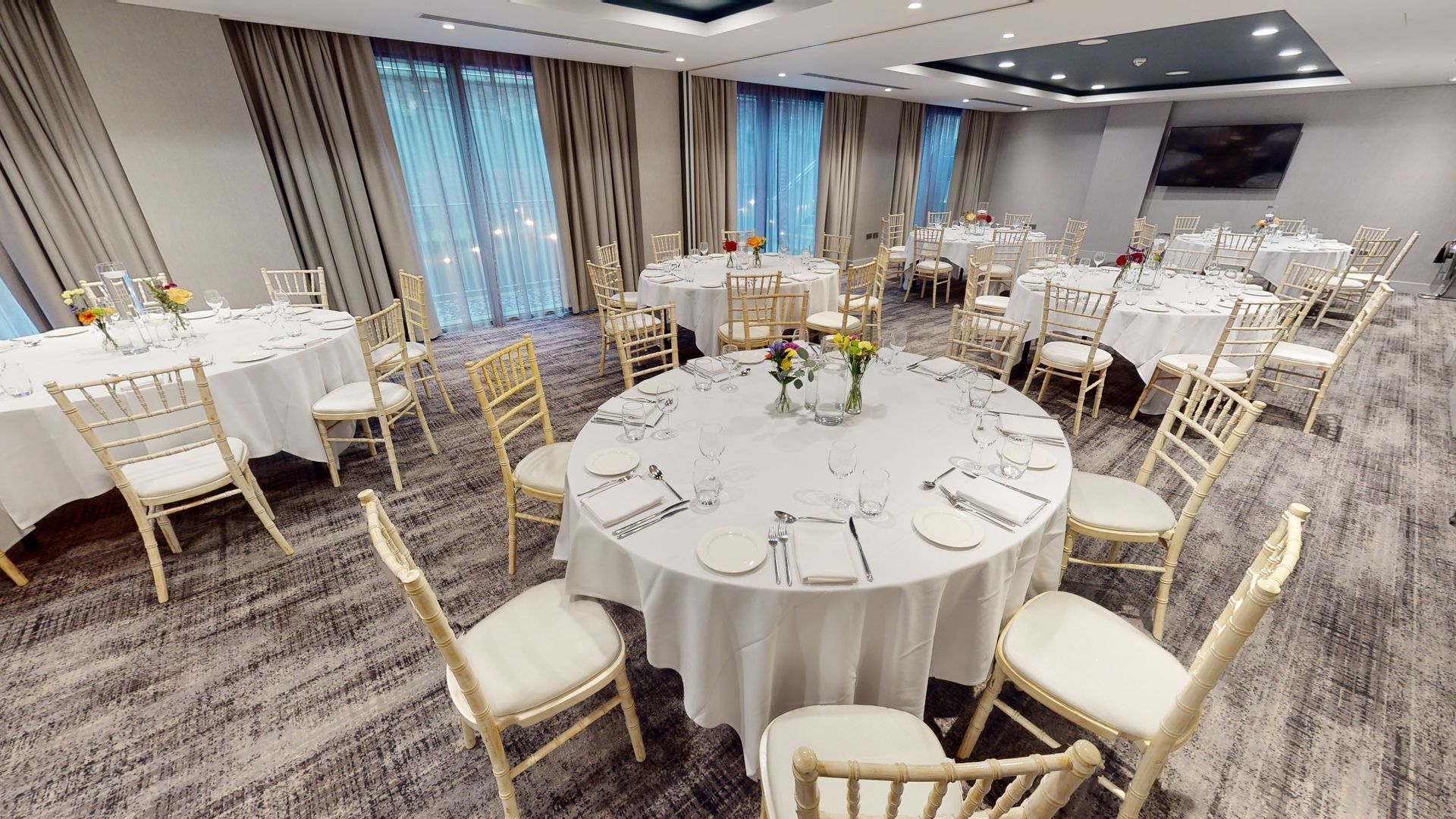 Banquet hall with round tables set for a meal, cream chairs, and large windows.