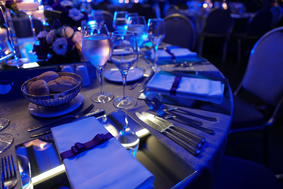 Elegant table setting with silverware, folded napkins, and bread in a silver bowl under blue lighting.