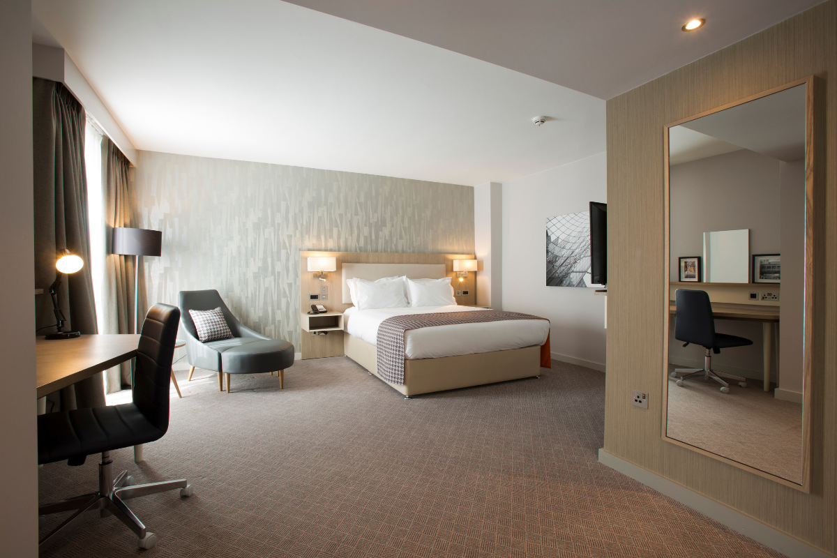 Hotel room with bed, chair, desk, large mirror; neutral tones.