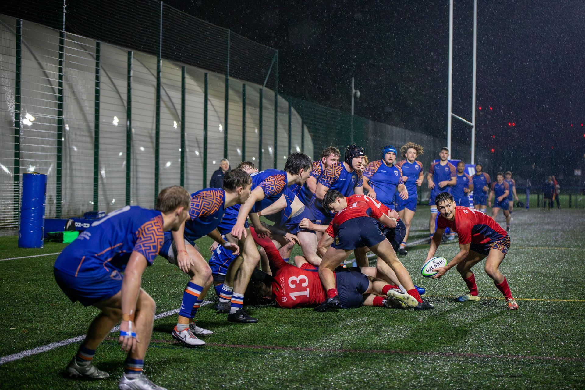 Rugby players in blue and red uniforms, scrumming on a green field under the lights on a rainy night.