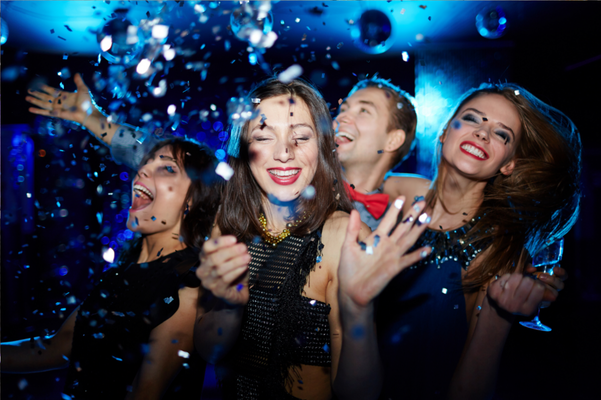 Four people laughing in a dark room with confetti; two women and a man with bright blue lighting.