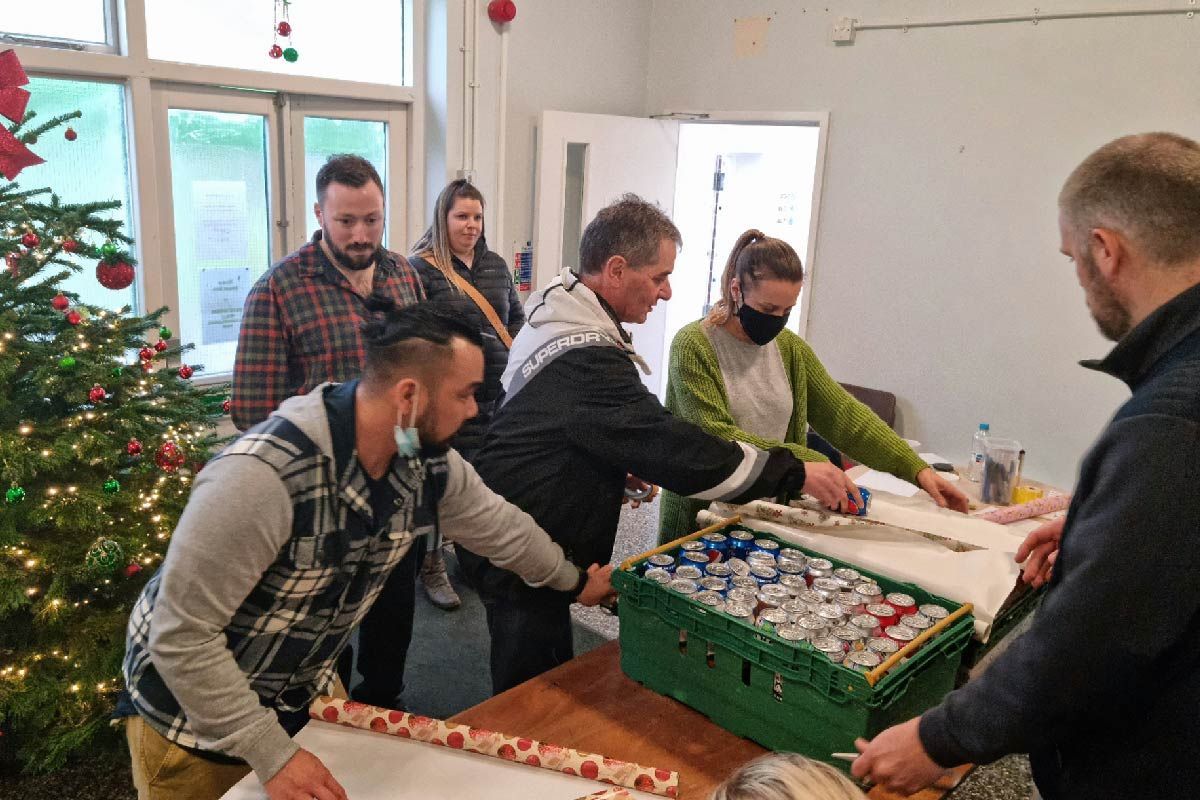 People wrapping gifts at a table with a Christmas tree. One wears a mask.