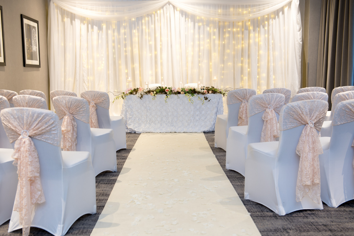 Wedding ceremony setting: white chairs with peach sashes face a decorated table and backdrop with a light carpet runner.