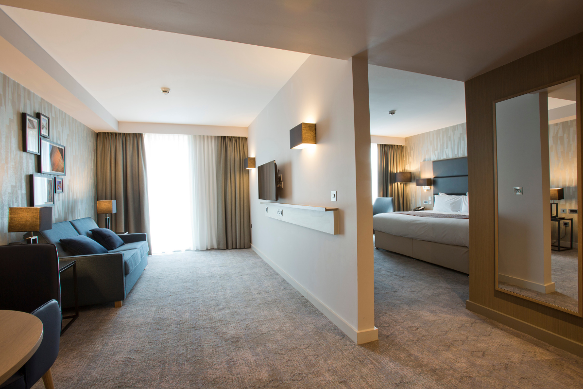 Hotel suite with a sitting area, bedroom, and large mirror. Gray and beige color scheme.