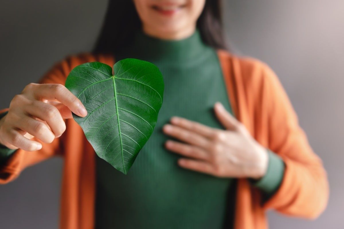 Woman holds green leaf shaped like a heart, hand on chest, smiling, wearing orange cardigan.