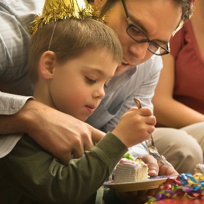 A boy in a party hat cuts cake with help from a man, possibly at a birthday party.