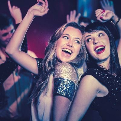 Two women dancing with joy at a party, surrounded by other people in a dimly lit setting.