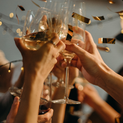 People toasting with wine glasses. Gold confetti falls, blurred lights in background.
