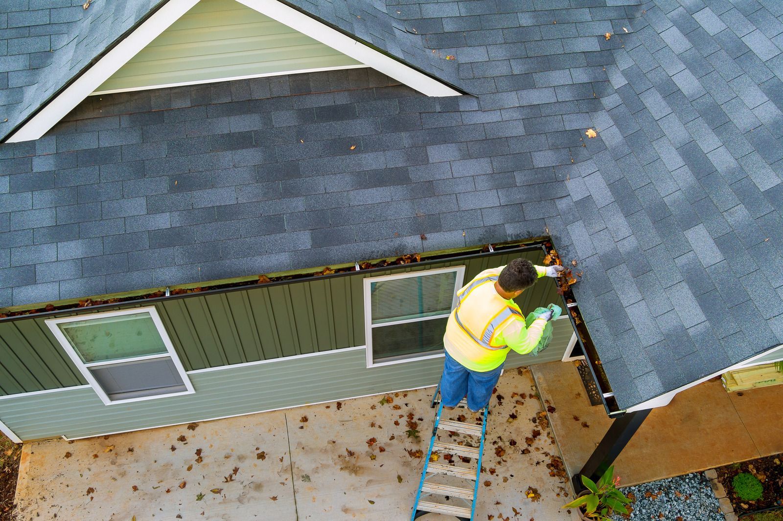Worker on ladder repairing roof edge of a house with gray shingles.