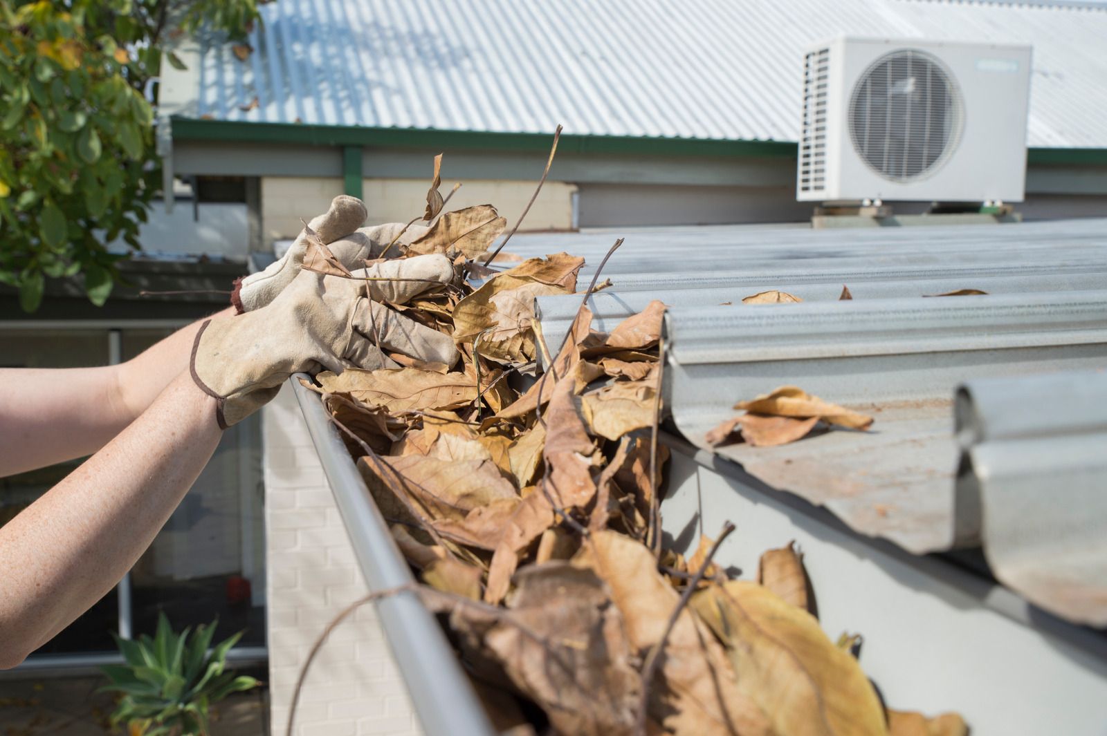 Hands clearing dry leaves from a clogged metal roof gutter near an air conditioner unit