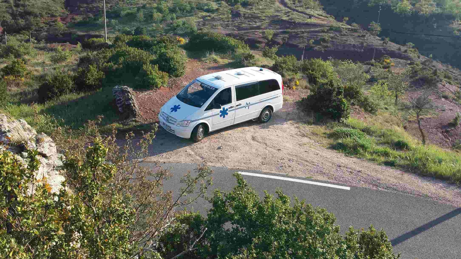 Une camionnette blanche est garée au bord d'une route de gravier, près de buissons verdoyants et de flancs de collines rocheuses.