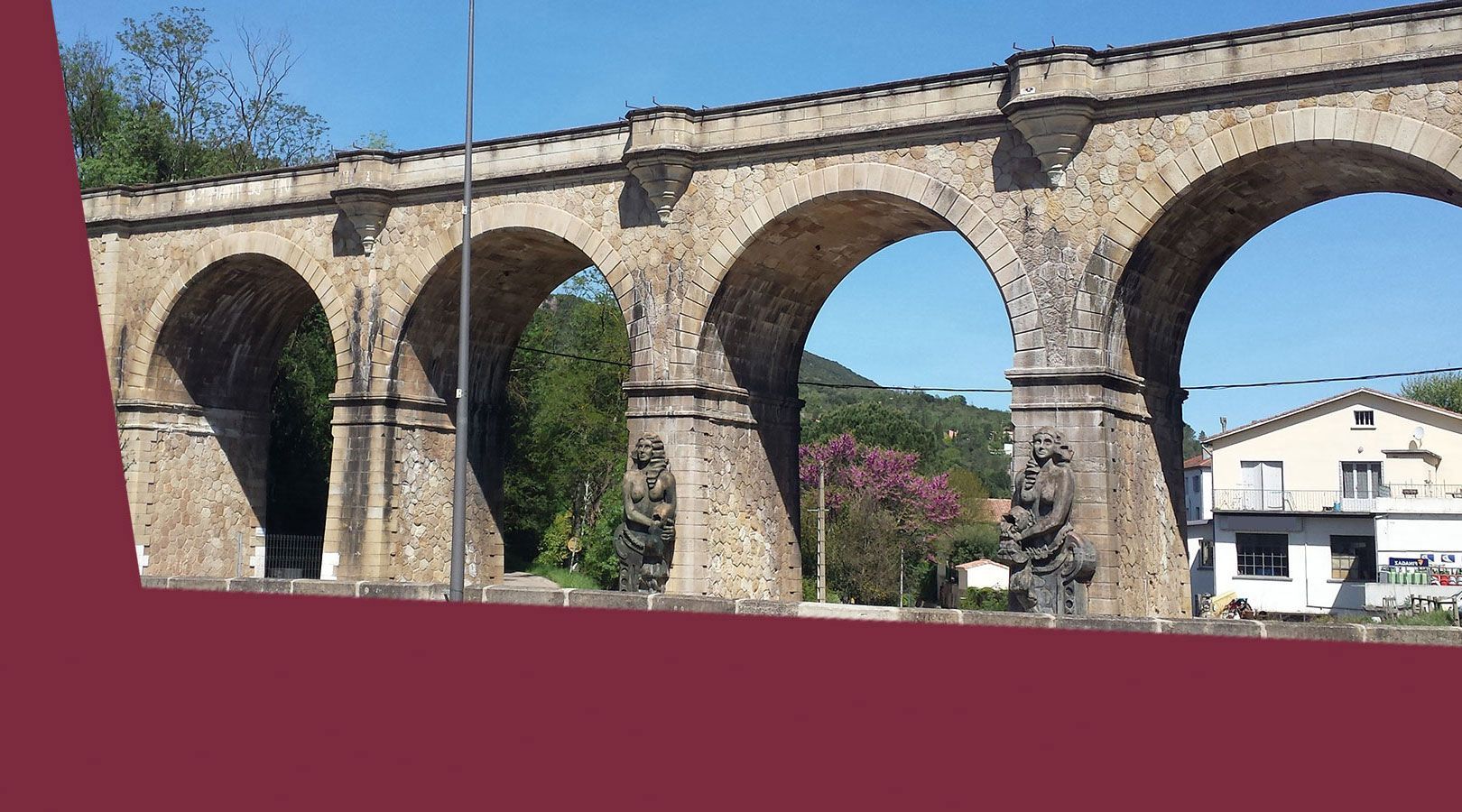 Aqueduc en pierre aux grandes arches surplombant un village, se détachant sur un ciel bleu et des arbres.