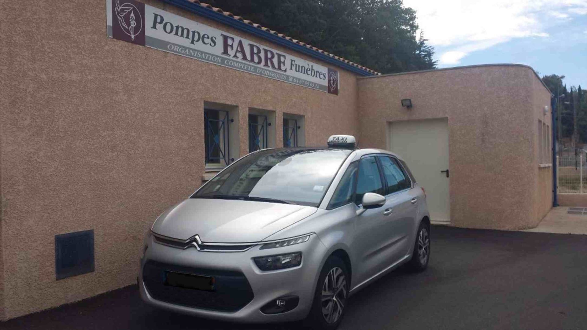 Une Citroën argentée garée devant un bâtiment avec un panneau Pompes Funèbres.