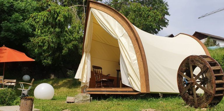 Zeltartige Glamping-Unterkunft mit cremefarbenem Vordach und Holzterrasse in einem grünen Garten