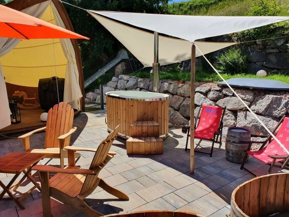 Terrassenbereich mit Sonnensegeln, Tischen aus Holzfässern und roten Klappstühlen neben einer Steinmauer