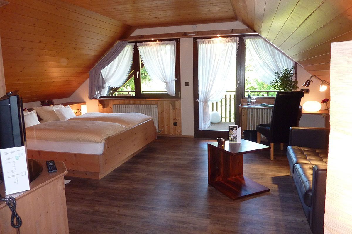 Dachzimmer mit Holzvertäfelung, Bett, Schreibtisch, Stuhl und großen Fenstern mit Blick auf einen Balkon.