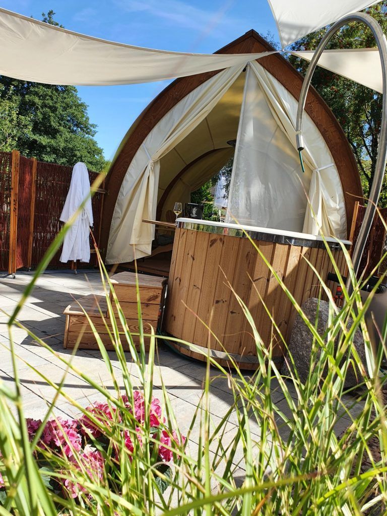 Outdoor-Glampingzelt mit weißen Sonnensegeln, Holzterrasse und rosa Blumen im Vordergrund