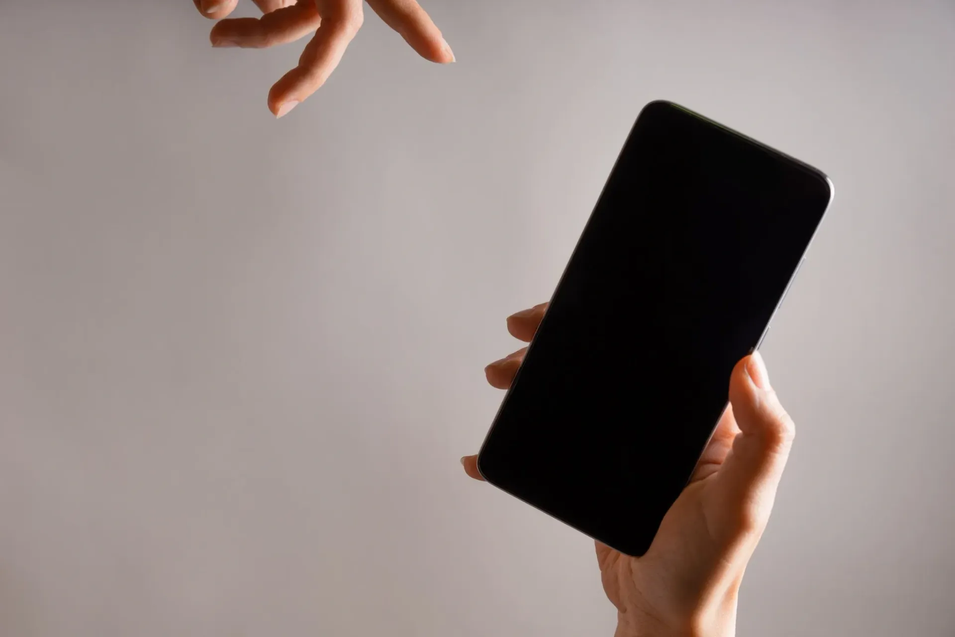Mano sosteniendo un teléfono inteligente negro con la otra mano apuntando hacia él sobre un fondo blanco.