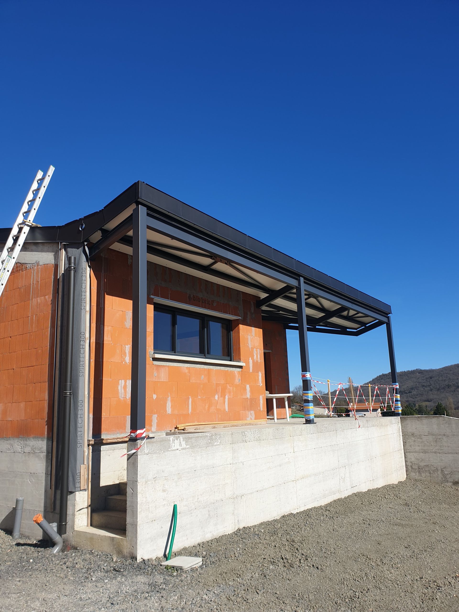 Petite maison en construction avec auvent métallique, murs de fondation blancs et cour gravillonnée.