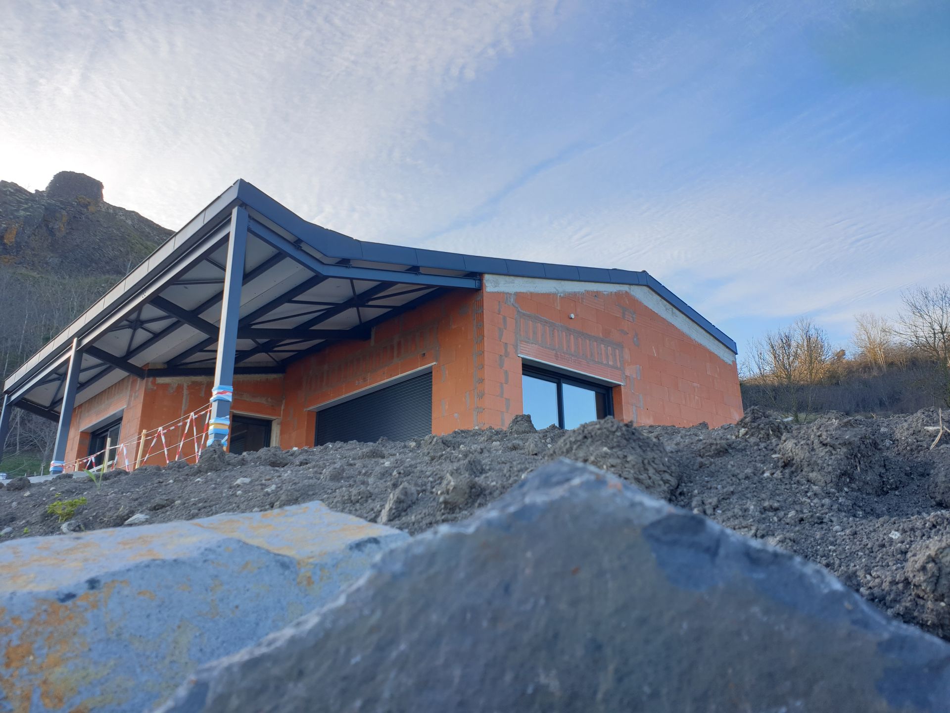 Maison moderne au toit bleu et aux murs orange, perchée sur un flanc de colline rocailleux.