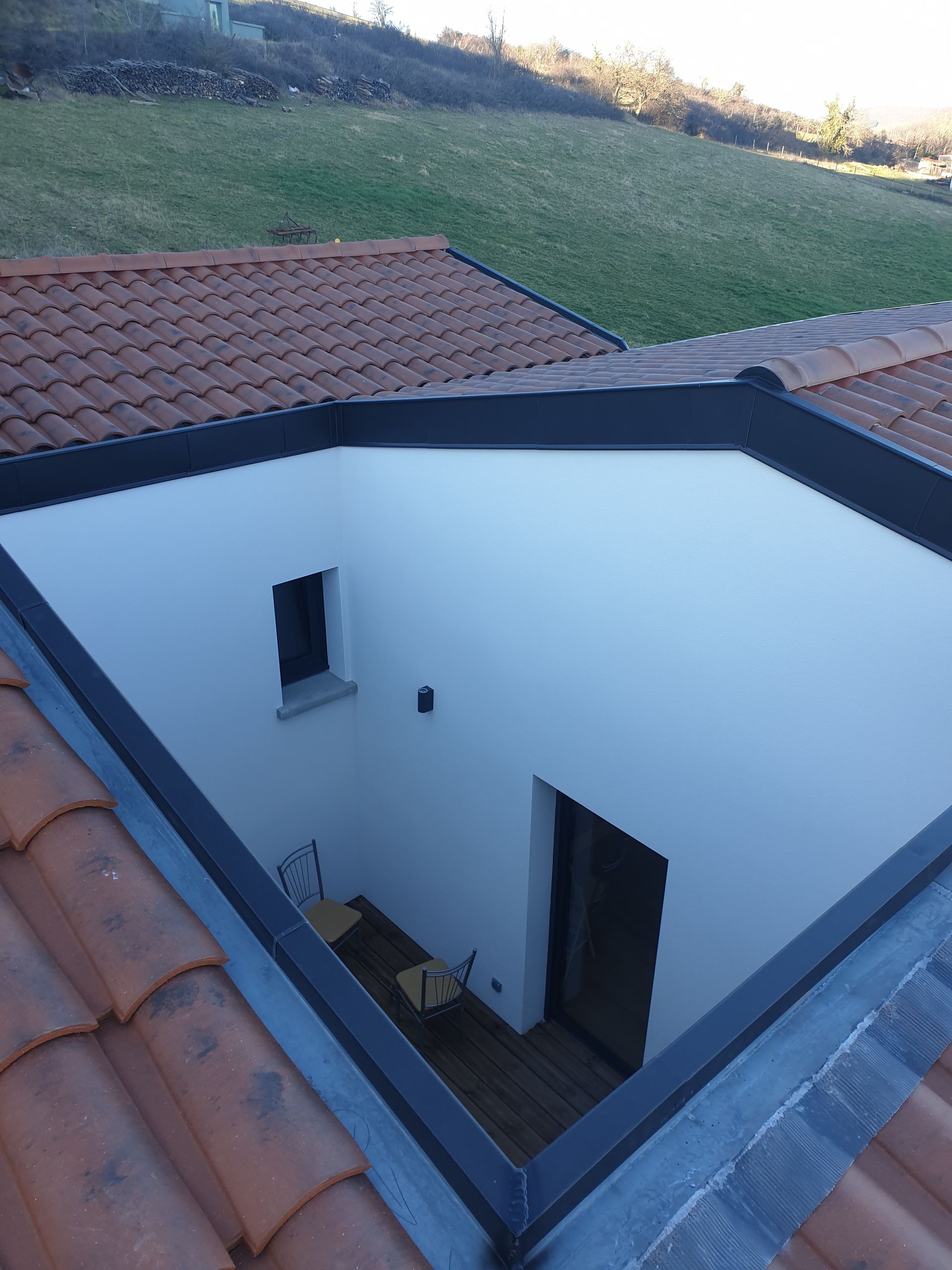 Vue plongeante sur une cour blanche aux boiseries noires et aux toits de tuiles rouges, à côté d'un talus herbeux.