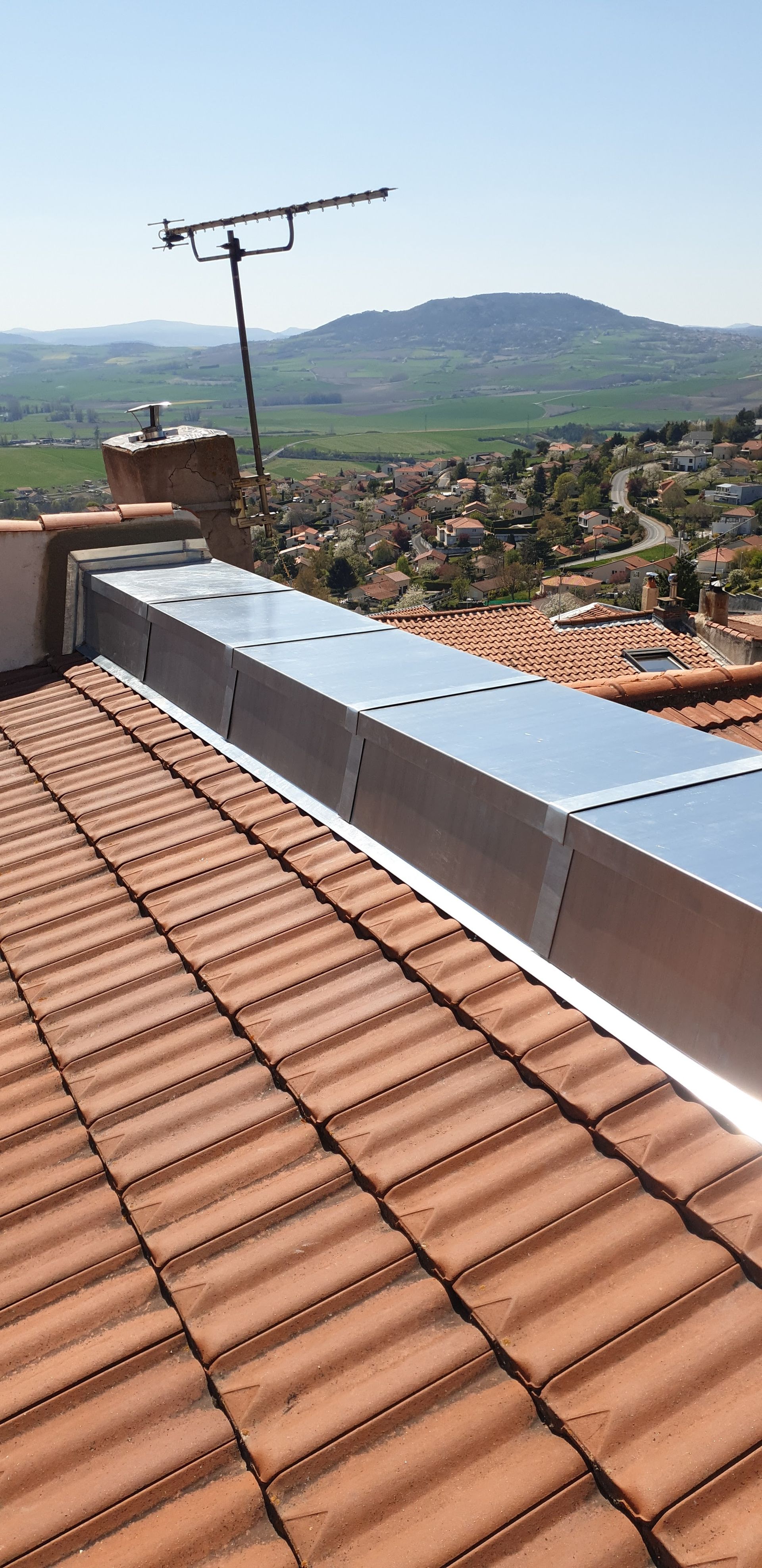 Aérateur métallique sur un toit de tuiles rouges donnant sur une ville à flanc de colline.