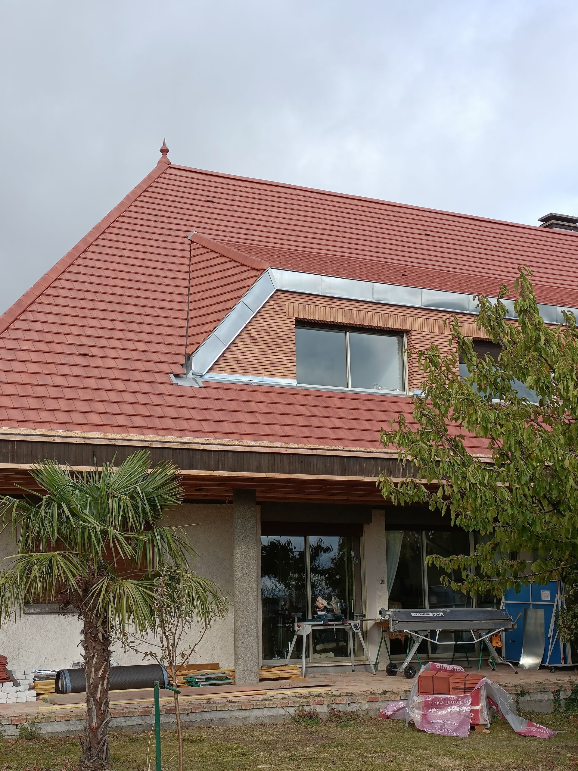 Maison au toit de tuiles rouges et lucarne inachevée, avec des matériaux de construction devant l'entrée.