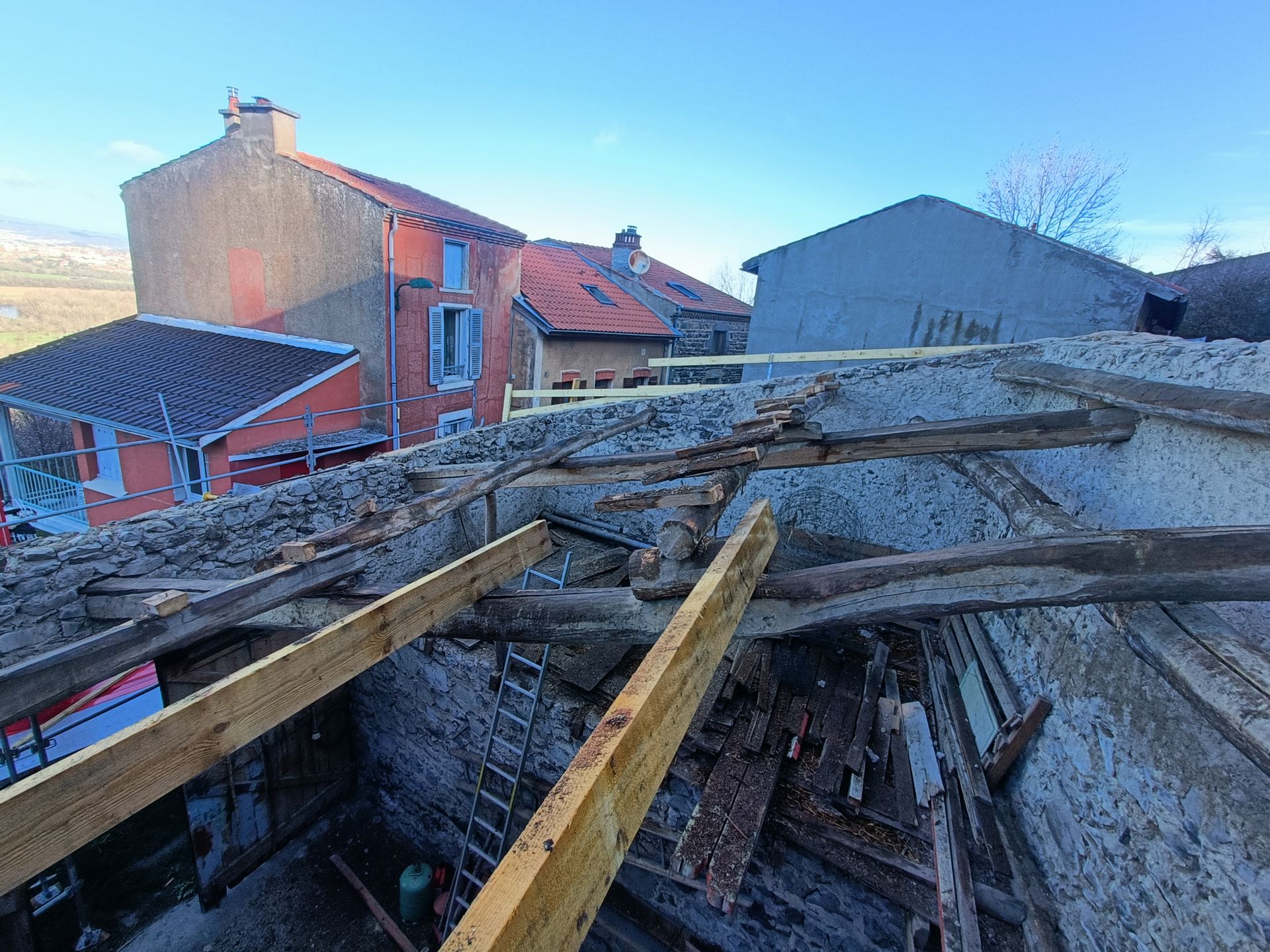 Des décombres et des poutres de bois brisées jonchent un toit effondré, à côté de maisons rouge et grise.