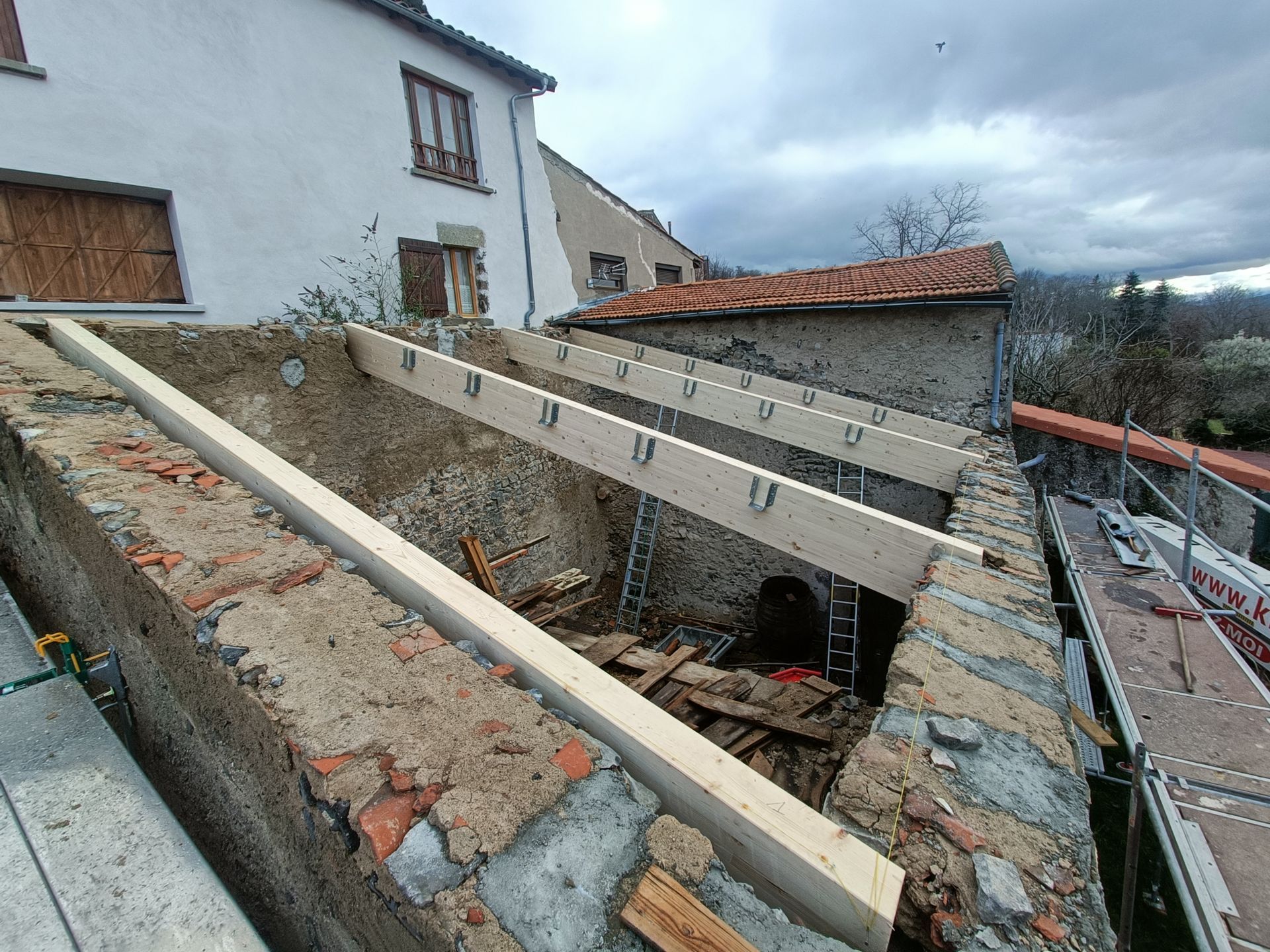 Toit endommagé avec poutres en bois apparentes et gravats à côté d'une maison blanche.