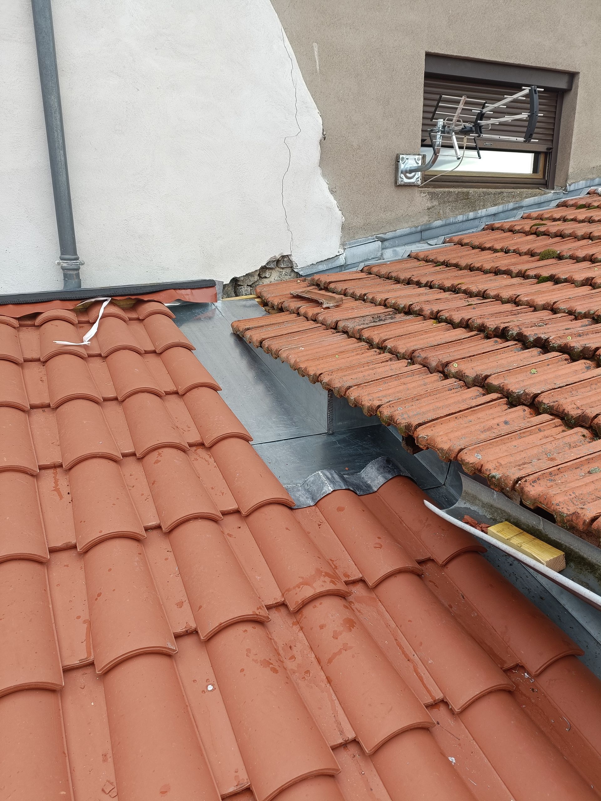 Des tuiles de toit se rejoignent le long d'un toit en pente, près d'un mur et d'une fenêtre, avec un joint d'étanchéité gris.
