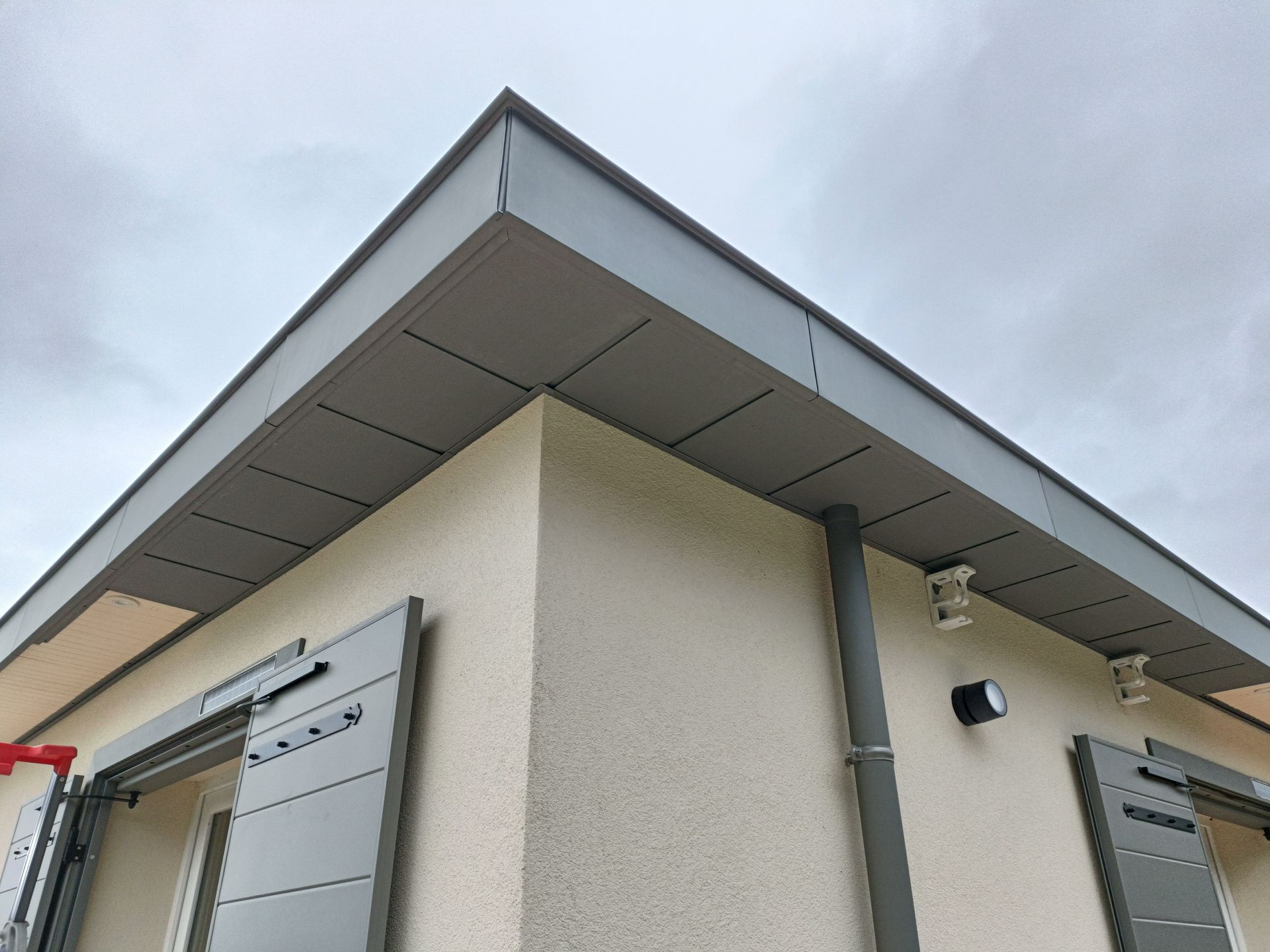 Coin d'un bâtiment beige au toit gris et aux luminaires muraux, sous un ciel couvert.