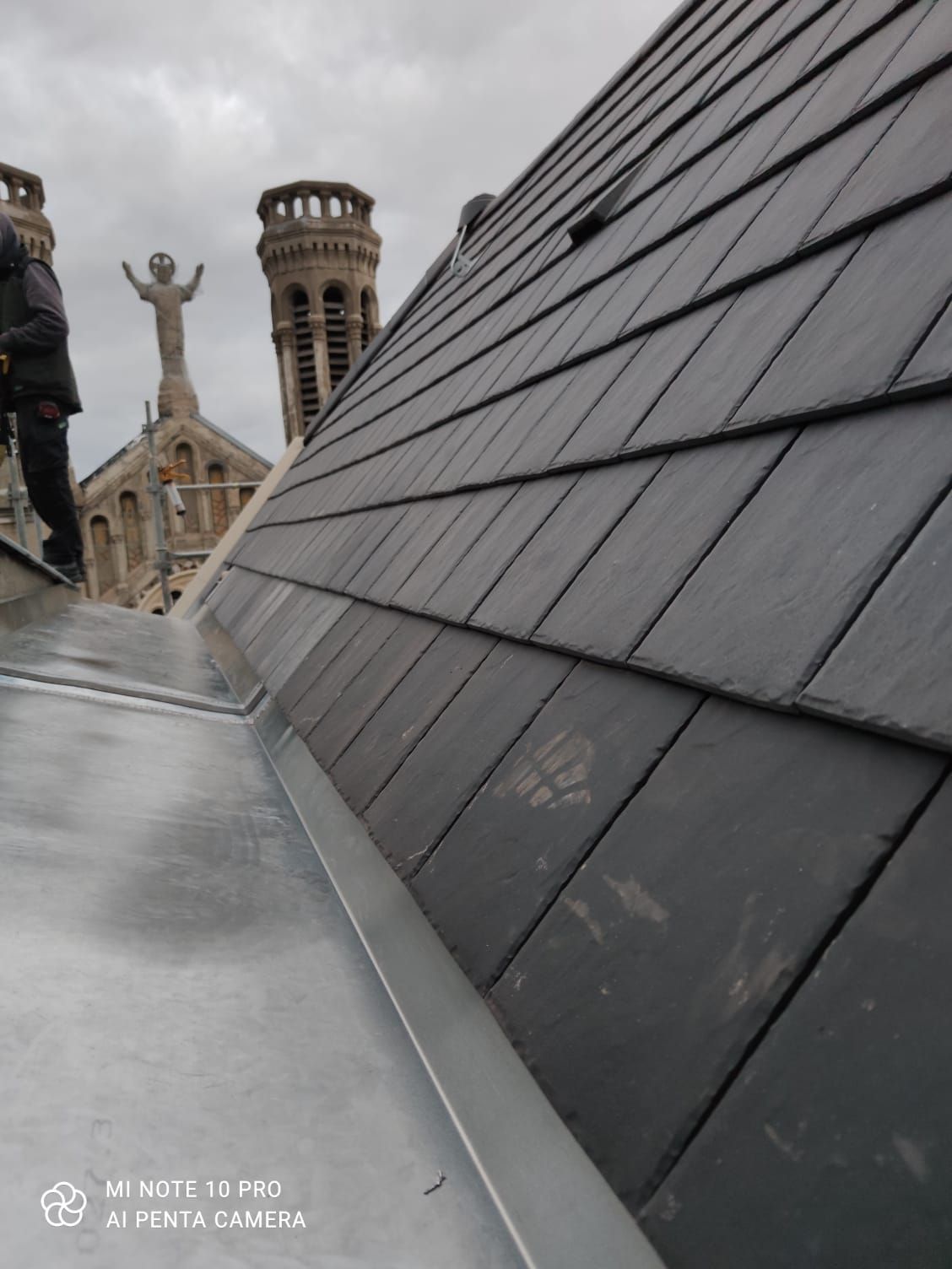 Toit en pente d'ardoise grise avec des bâtiments de la ville et une tour visibles en arrière-plan.